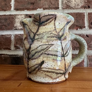 May include: A handmade ceramic pitcher with a leaf design. The pitcher is beige with brown and green leaf patterns. It has a curved handle and a spout. The pitcher is sitting on a wooden surface in front of a brick wall.
