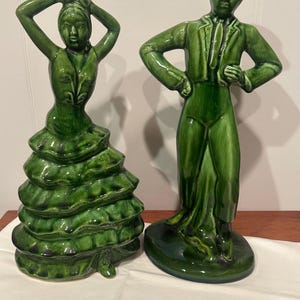 May include: Two green ceramic figurines: a woman in a ruffled dress with arms raised, and a man in a suit and hat. The figurines are glossy and stand on a wooden surface.