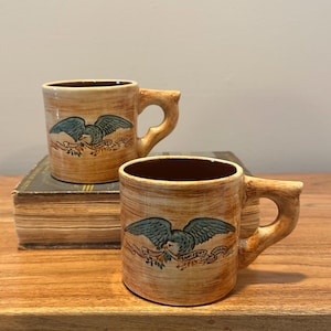 May include: Two vintage-style ceramic mugs with a tan, textured finish. Each mug features a teal eagle emblem with a banner. The mugs have brown interiors and unique, branch-like handles. They are displayed on a wooden surface.