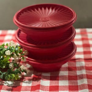 May include: A stack of three round, red plastic storage containers with matching lids. Each lid features a distinct sunburst or starburst pattern radiating from the center. The containers are uniform in color and design, stacked vertically.