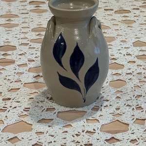 May include: A beige ceramic vase with a rounded body and a small opening. The vase features two small handles and a blue leaf design. The vase is sitting on a white lace doily.