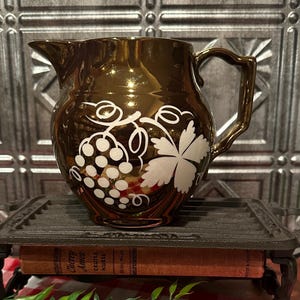 May include: A shiny, bronze-colored pitcher with a white grape and leaf design. The pitcher has a handle and spout. It sits on a decorative metal stand with a red and white checkered tablecloth in the background.