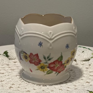 May include: A white ceramic bowl with a scalloped rim and a gold-tone edge. The exterior features a floral pattern with red and yellow flowers, green leaves, and small blue accents. The bowl has a raised, embossed decorative line near the top.