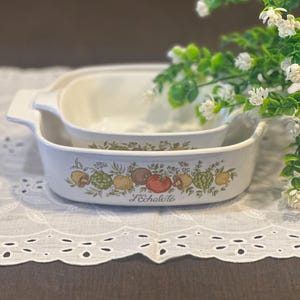 May include: Two white ceramic baking dishes with integrated handles, featuring a colorful botanical illustration of vegetables and the text L'Echalote printed on the front of the larger dish, nested one inside the other.