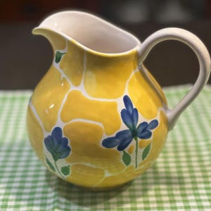 May include: A yellow ceramic pitcher featuring a white abstract pattern and hand-painted blue flowers with green leaves. The pitcher has a curved handle and a wide pouring spout, with a smooth, glossy finish throughout the exterior surface.