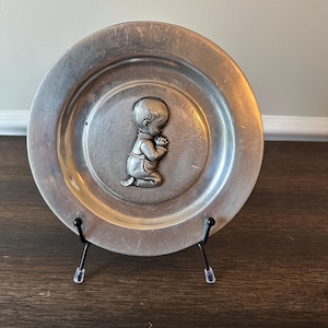 May include: A small, round, silver-colored plate with a raised relief of a kneeling baby in prayer. The plate is displayed on a black metal stand. The plate has a slightly worn appearance.