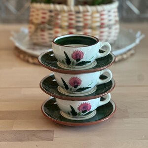 May include: A stack of three ceramic teacups and saucers. The cups are white with a green interior and a pink thistle design. The saucers are green with a brown rim. The cups are stacked on top of each other.