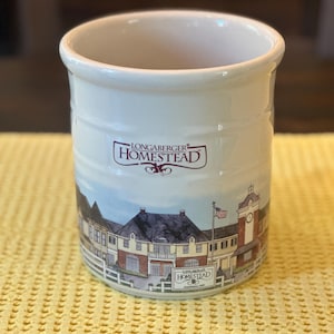 May include: A cylindrical cream-colored ceramic crock featuring the Longaberger Homestead logo in dark red text. The exterior displays a detailed illustration of a building complex with a clock tower, a white fence, and an American flag.