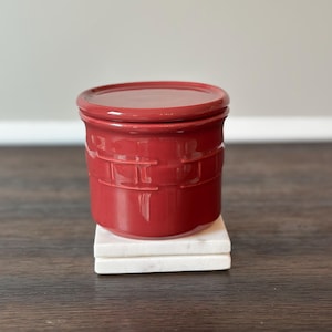 May include: A small, round, red ceramic jar with a matching lid. The jar has a textured, basket-weave design around the middle and sits on a stack of two white marble coasters. The background is a neutral gray wall.