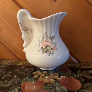 May include: White ceramic pitcher with a curved handle and floral design. The pitcher features pink and green floral patterns on the front and handle. The pitcher has a ribbed texture and a wide spout.