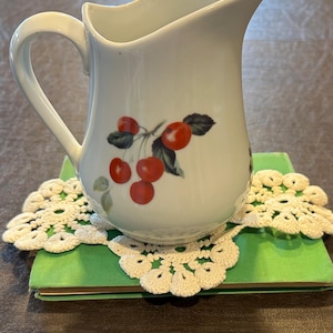 May include: White ceramic pitcher with a curved handle and a cherry design. The pitcher is resting on a green book with a cream-colored, crocheted doily. The cherries are red with green leaves.