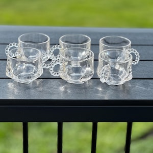 May include: Six clear glass teacups with decorative beaded handles. The cups have a faceted design on the lower portion and sit on a dark gray surface. The background is a blurred green.