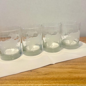 May include: Four clear, cylindrical drinking glasses with a frosted world map design. The glasses are arranged on a white cloth, set against a light background. The glasses have a grid pattern and a frosted world map design.