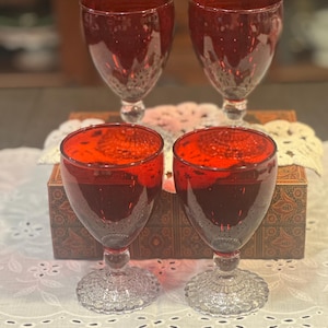 May include: Four deep red glass goblets with clear, textured, circular bases. Each goblet features a smooth, rounded bowl and a short, thick stem that connects to a wide, patterned foot with a beaded edge, creating a uniform set of glassware.
