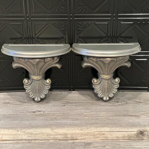 May include: A pair of matching semi-circular wall shelves featuring a metallic silver finish and ornate, carved-style supports with decorative scrollwork details. Each shelf has a flat top surface and a tapered base with a central vertical design.