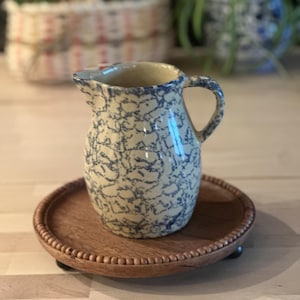 May include: A ceramic pitcher with a blue and white pattern sits on a round wooden tray. The pitcher has a handle and a spout. The tray has a beaded edge and small feet. The background is blurred.