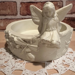 May include: Off-white ceramic fairy bowl with a seated fairy figure and dragonfly and leaf relief details. The bowl has a round shape and is placed on a white lace doily. The background is a brick wall.