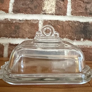 May include: Clear glass butter dish with a domed lid and decorative edges. The lid features a circular handle. The dish is rectangular and sits on a wooden surface, with a brick background.
