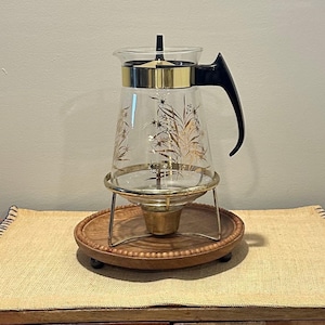 May include: A vintage glass coffee pot with a gold rim and black handle. The clear glass is decorated with gold floral designs. It sits on a gold metal stand and a wooden base.