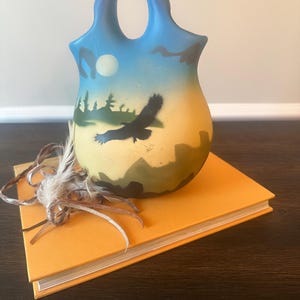 May include: A hand-painted ceramic vase with a handle, featuring a landscape scene with an eagle in flight. The vase is set on an orange book, with feathers tied around the spine. The vase has a blue top, a yellow middle, and a green and black landscape design.