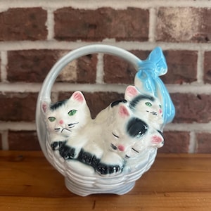 May include: A ceramic basket-shaped figurine featuring three white cats with black markings and green eyes. The cats are nestled together in a woven basket with a handle. A blue bow is attached to the handle. The figurine is set against a brick wall.