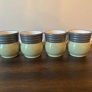 May include: Four ceramic mugs with a green base and a gray, ribbed top. Each mug has a white interior and a curved handle. The mugs are arranged in a row on a dark wooden surface.