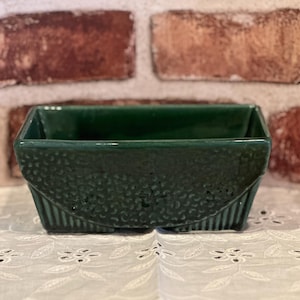 May include: A rectangular, emerald green ceramic planter. The planter has a textured, semi-circular design on the front and vertical lines on the sides. The planter is sitting on a white, embroidered surface.
