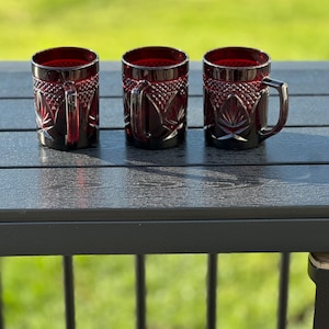 May include: Three ruby red glass mugs with a diamond and leaf pattern. Each mug has a handle and a thick base. The mugs are displayed on a dark gray surface with a green background.