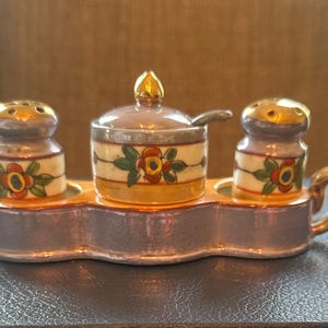 May include: A vintage ceramic salt and pepper set with a matching lidded container and spoon. The set features a floral design with gold accents on a gray base. The set includes two salt shakers and a lidded container, all resting on a tray with handles.