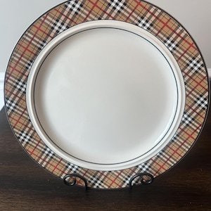 May include: A white ceramic plate with a brown, red, and black plaid border. The plate is displayed on a black metal stand, showcasing a classic design suitable for dining or display.