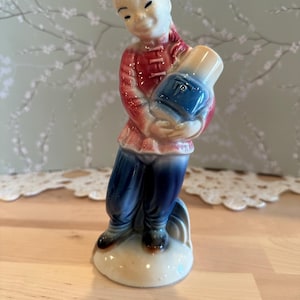 May include: A vintage ceramic figurine of a smiling Asian figure in traditional attire, holding a blue and beige vase. The figurine is painted with red, blue, and beige colors. The figure is standing on a beige base.