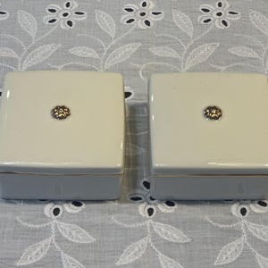 May include: Two square, off-white ceramic trinket boxes with smooth, glossy finishes. Each lid features a small, centered, metallic floral-shaped knob. The boxes have a simple, clean design with thin gold-toned trim along the edges of the lids.