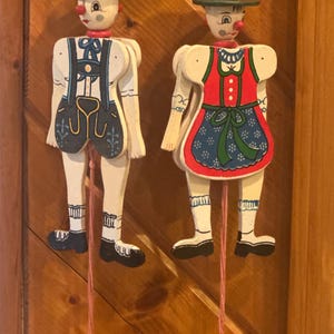 May include: Two wooden marionette dolls, one dressed in blue lederhosen with suspenders and the other in a red and blue dress. Both dolls have painted faces, green hats, and red noses. Each doll has a string with a red bead at the end.