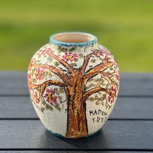 May include: A round ceramic vase with a hand-painted tree design. The tree has brown branches and is adorned with pink and red flowers. The vase has a cream-colored background and a turquoise rim. The text "KAPOK TREE" is visible on the vase.