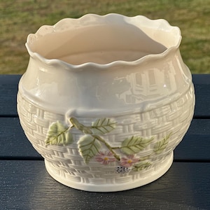 May include: A cream-colored ceramic planter with a scalloped rim and a textured basket-weave pattern on the exterior. The surface features a raised, three-dimensional relief of a branch with green leaves, small pink flowers, and a dark berry.