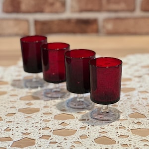 May include: Four ruby red wine glasses with clear stems are arranged on a cream-colored, open-weave lace tablecloth. The glasses are cylindrical with a slight taper and are evenly spaced.