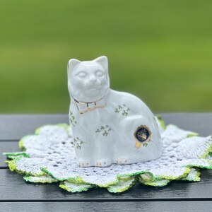 May include: A white porcelain cat figurine featuring a gold-trimmed collar and small green floral patterns on its body. The cat is sitting on a round, white crocheted doily with a green-tinted scalloped edge, displaying a gold circular sticker.