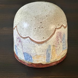 May include: A dome-shaped ceramic object with a speckled off-white top and a scalloped edge. The lower section features a hand-painted landscape design with vertical blue, yellow, and purple shapes against a cream background, finished with a reddish-brown base.
