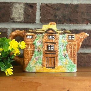 May include: A ceramic teapot shaped like a house, featuring a brown roof and walls with painted windows and a door. The teapot has a handle and spout, and is decorated with green and pink floral accents. The teapot is sitting on a wooden surface.