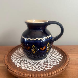 May include: A small, dark blue ceramic pitcher with a handle, decorated with yellow and orange floral patterns. The pitcher sits on a wooden tray with a white lace doily. The tray has a beaded edge.