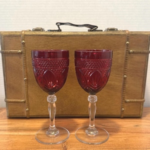 May include: Two ruby red crystal wine glasses with intricate diamond and leaf patterns. The glasses have clear stems and bases. They are set on a wooden surface, with a vintage wooden suitcase in the background.