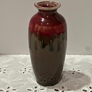 May include: A ceramic vase with a glossy finish. The top is a deep red, transitioning to a brown base with a dripped glaze effect. The vase has a narrow neck and a rounded body, suitable for displaying flowers.