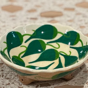 May include: A small, ceramic bowl with a scalloped rim and three small feet. The bowl is painted with a cream-colored interior and a pattern of green chili peppers. The exterior is tan with green accents.