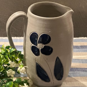 May include: A light gray ceramic pitcher featuring a single handle and a spout. The exterior is decorated with a dark blue floral design consisting of a five-petaled flower and two elongated leaves, all rendered in a smooth, glossy finish.