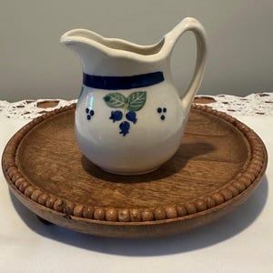 May include: A small, white ceramic pitcher with a curved handle and a spout, decorated with blue and green blueberry motifs. The pitcher sits on a round, wooden tray with a beaded edge. The tray is a warm brown color.