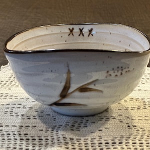 May include: A ceramic bowl with a light gray, speckled glaze and a dark brown rim. The exterior features a painted brown bamboo stalk design. The interior rim is decorated with three dark brown X symbols spaced evenly along the top edge.