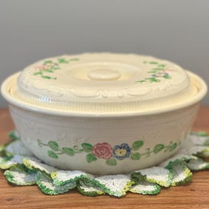 May include: A cream-colored ceramic casserole dish with a matching lid, featuring embossed decorative borders and a hand-painted floral pattern of pink roses, blue flowers, and green leaves around the center and on the lid surface.