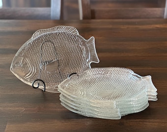 May include: A set of clear glass plates shaped like fish, featuring a textured scale pattern on the surface. One plate is displayed upright on a black metal stand, while a stack of four identical plates rests horizontally in front.