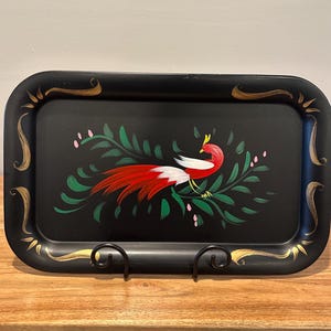 May include: Black rectangular serving tray with a hand-painted bird design. The bird has red and white feathers, a yellow crown, and is surrounded by green leaves and pink flowers. Gold accents adorn the tray's edges.