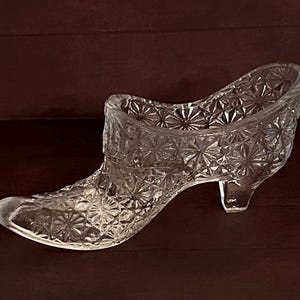 May include: A clear glass shoe with a detailed starburst pattern. The decorative shoe has a pointed toe and a small heel. The glass shoe is likely a decorative item or a small container.